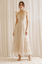 A woman wearing a beige maxi dress with embroidered flowers, featuring a plunging neckline and ruffled straps.
