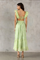 Back view of a woman wearing a lime green dress with ruffled sleeves and open back against a gray concrete wall.