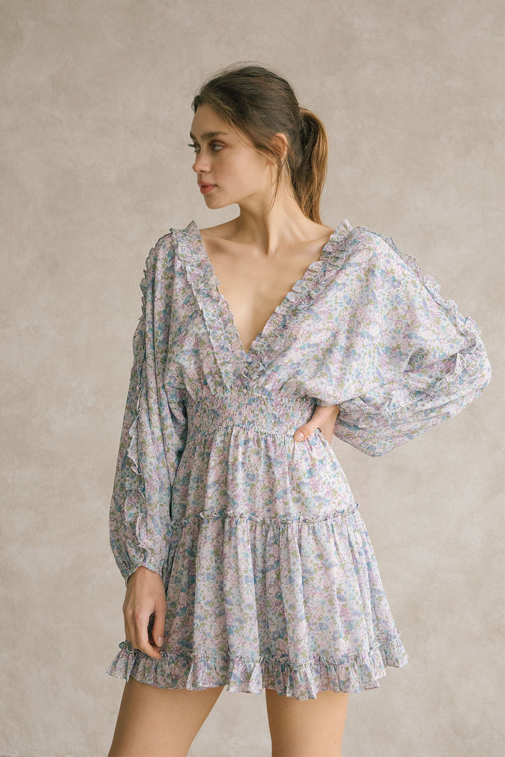 Woman wearing a blue dress with floral pattern and v-neckline with ruffle trimming against a neutral background