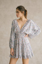 Woman wearing a blue dress with floral pattern and v-neckline with ruffle trimming against a neutral background