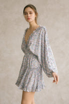 Side view of a woman wearing a blue dress with floral pattern and v-neckline with ruffle trimming against a neutral background