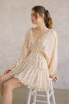 A model sitting on a white stool wearing a yellow dress with floral pattern against a gray wall
