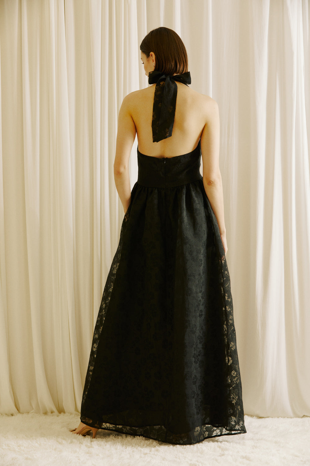 Back view of a woman wearing a black halter neck maxi dress against a beige curtain background
