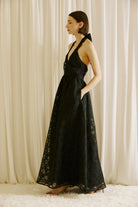 Side view of a woman wearing a black halter neck maxi dress against a beige curtain background