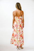 A woman viewed from behind wearing a sleeveless maxi dress with thin straps and a colorful floral print on a light cream background. She is standing against a plain white studio background.