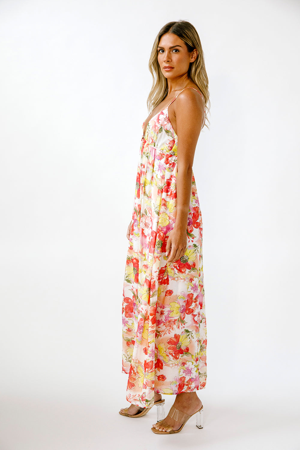 A woman modeling a sleeveless maxi dress with a vibrant red, yellow, and green floral print on a cream background, photographed against a plain white studio backdrop.