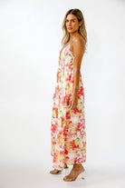 A woman modeling a sleeveless maxi dress with a vibrant red, yellow, and green floral print on a cream background, photographed against a plain white studio backdrop.