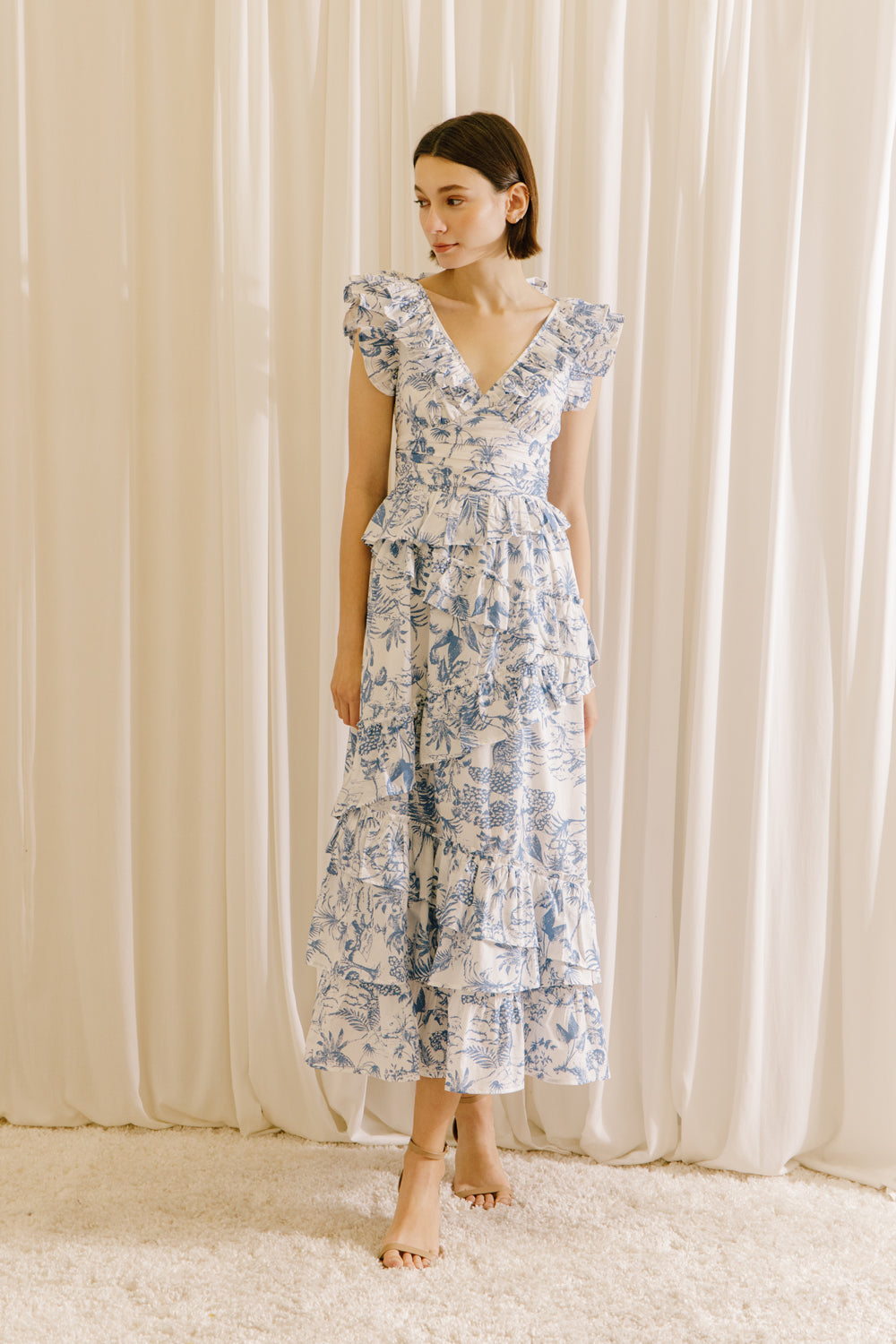 View of a woman wearing a white and blue tropical print midi dress against a beige curtain.