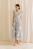 View of a woman wearing a white and blue tropical print midi dress against a beige curtain.