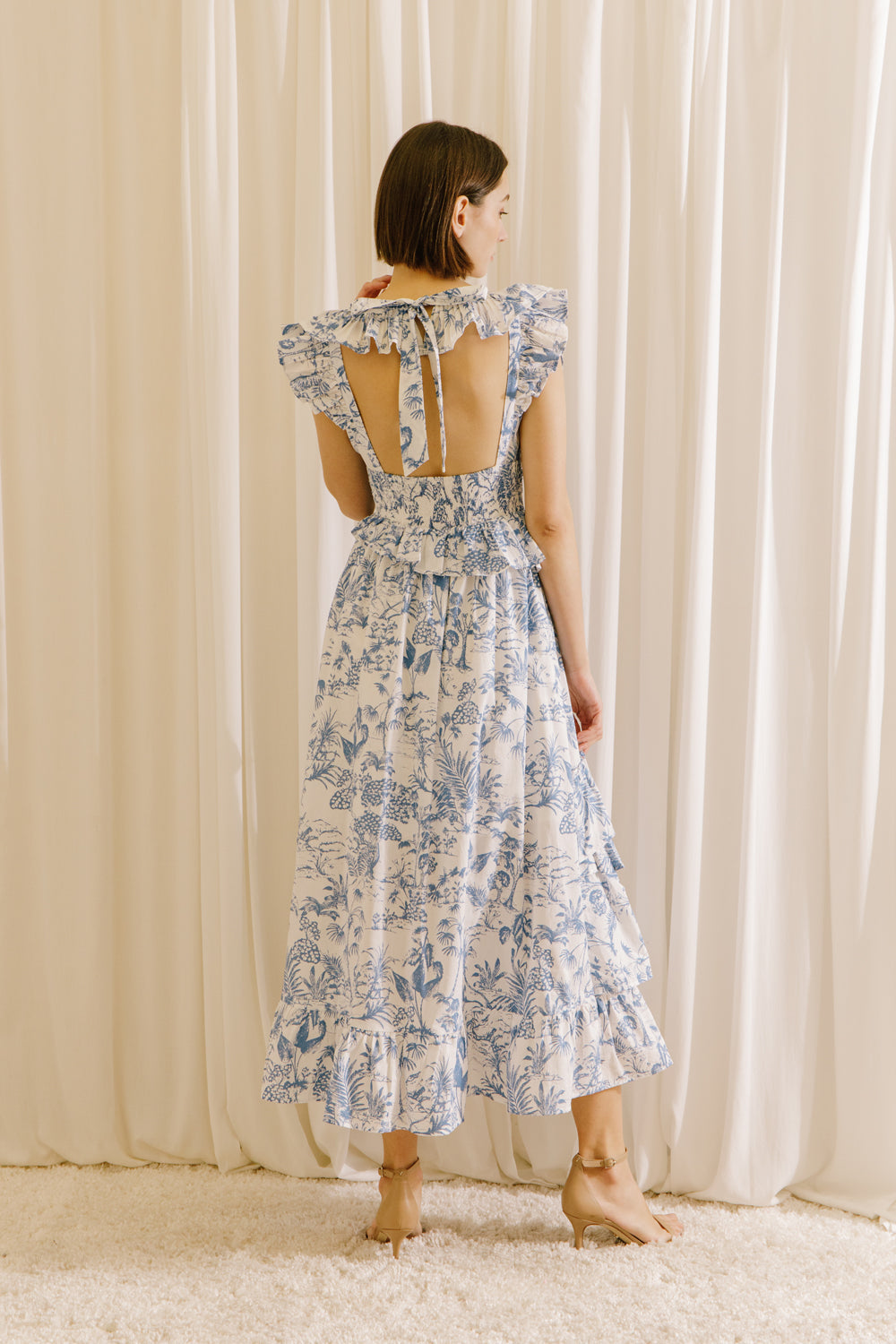 Back view of a woman wearing a white and blue tropical print midi dress standing against a beige curtain.