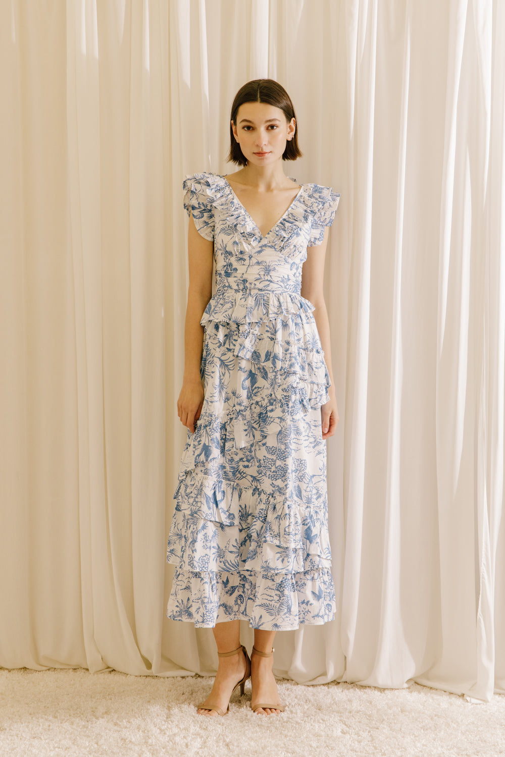 Woman wearing a white and blue tropical print midi dress standing against a beige curtain.