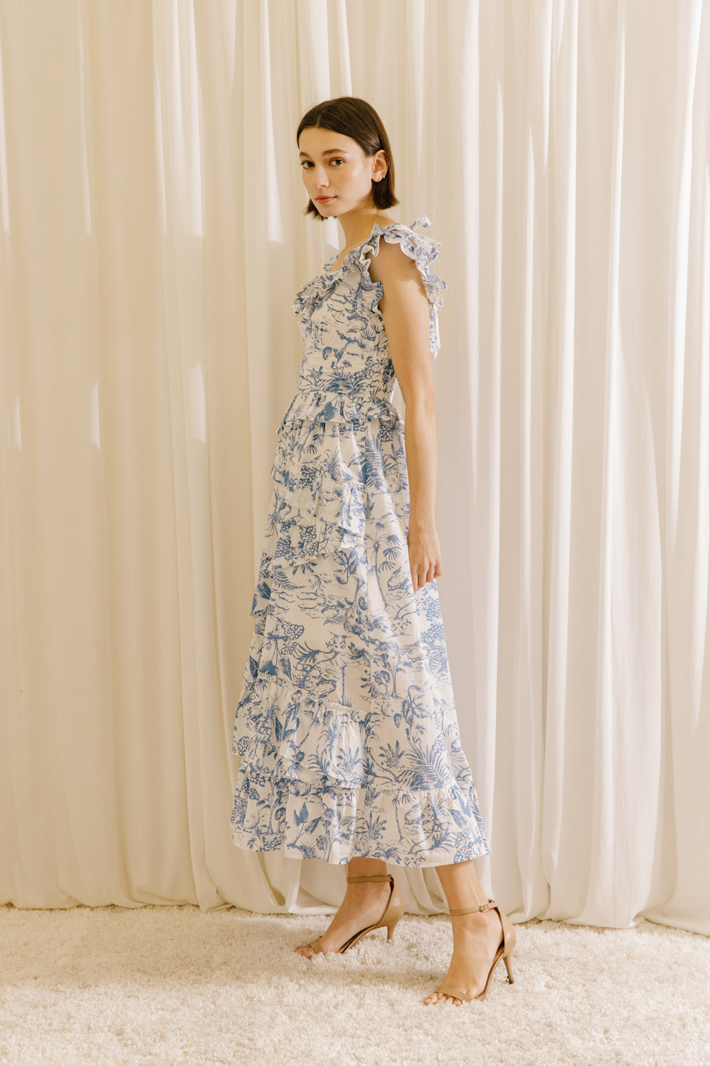 Side view of a woman wearing a white and blue tropical print midi dress standing against a beige curtain.