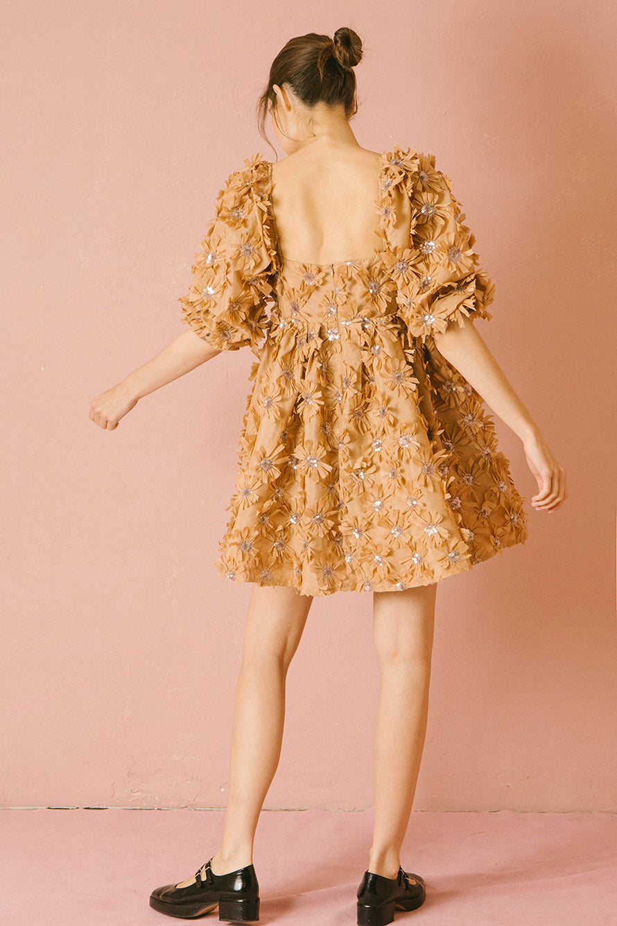 Back view of a woman wearing a caramel dress with 3D floral detail throughout against a pink background
