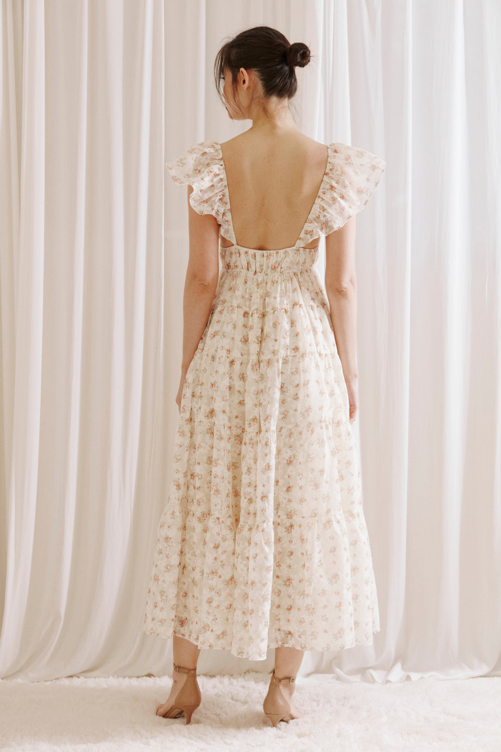 Back view of a woman wearing a floral maxi dress against a white curtain background