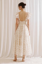Back view of a woman wearing a floral maxi dress against a white curtain background