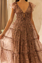 A person wearing a mauve midi dress with a floral pattern, flowy A-line silhouette, flutter sleeves and tiered skirt against a beige wall.