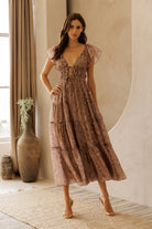 A model wearing a mauve midi dress with a floral pattern, flowy A-line silhouette, flutter sleeves and tiered skirt against a beige wall.