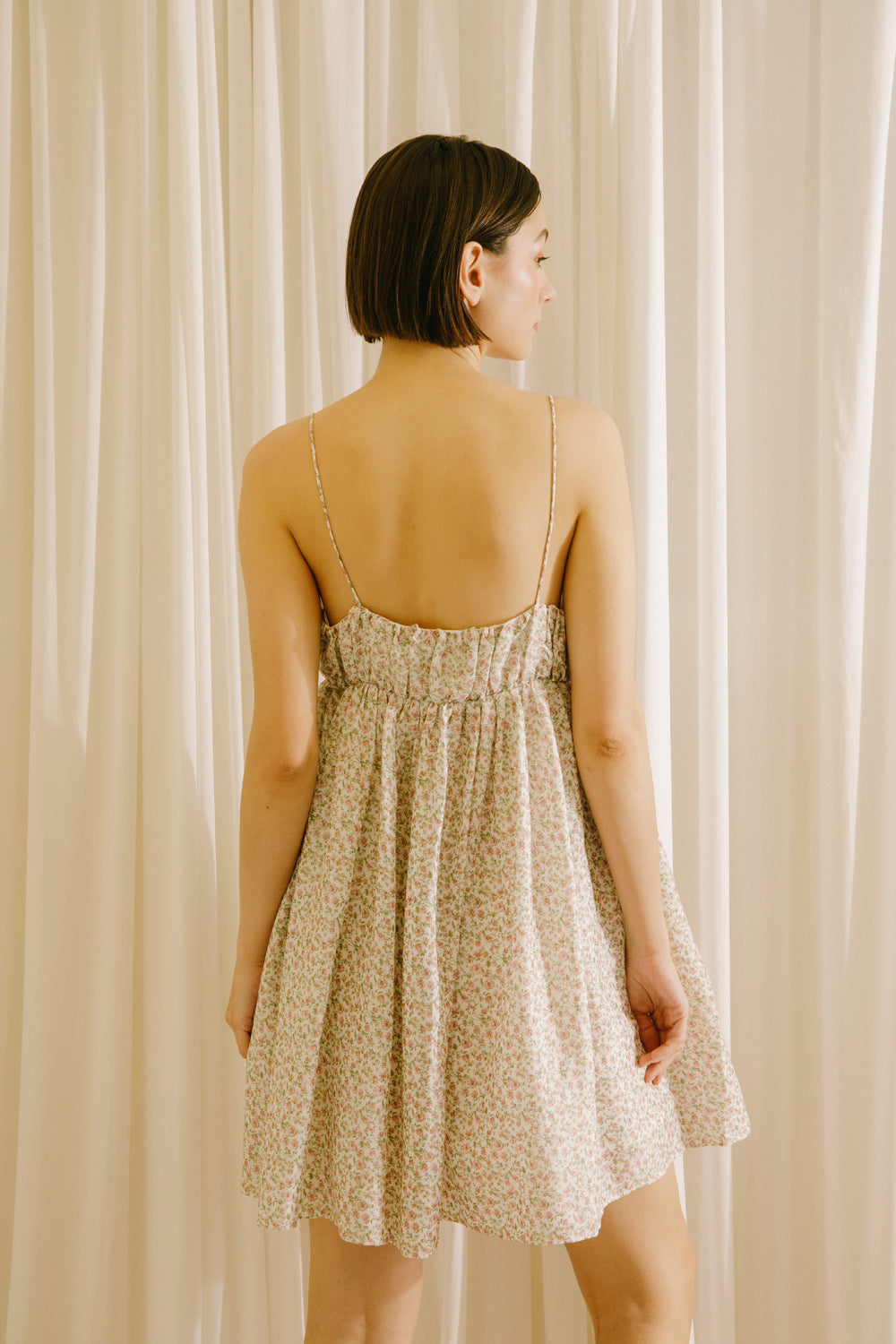 Back view of a woman wearing a beige mini dress against a beige curtain background