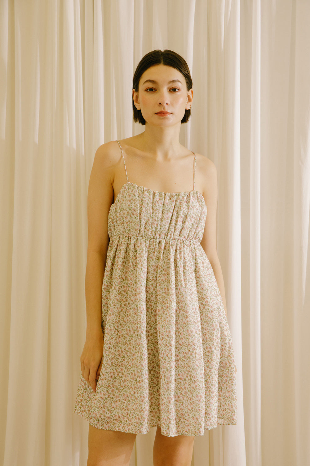 Woman wearing a beige dress with pink floral pattern standing against a beige curtain