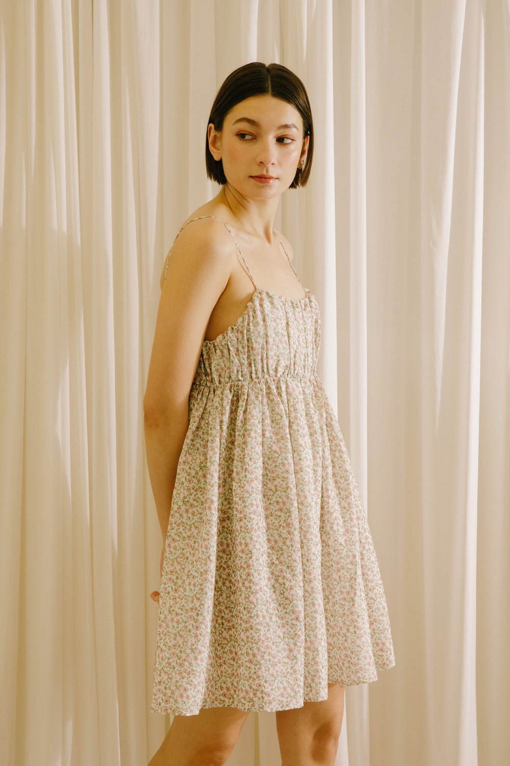 Side view of a woman wearing a beige mini dress against a beige curtain background