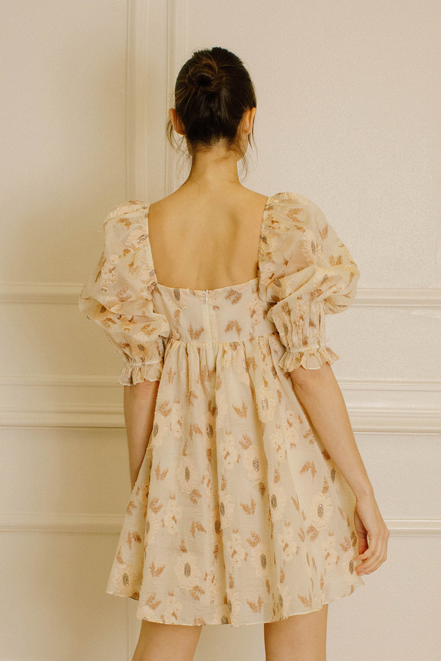 Back view of a woman wearing a beige dress with a floral pattern and an invisible back zipper in an indoor setting
