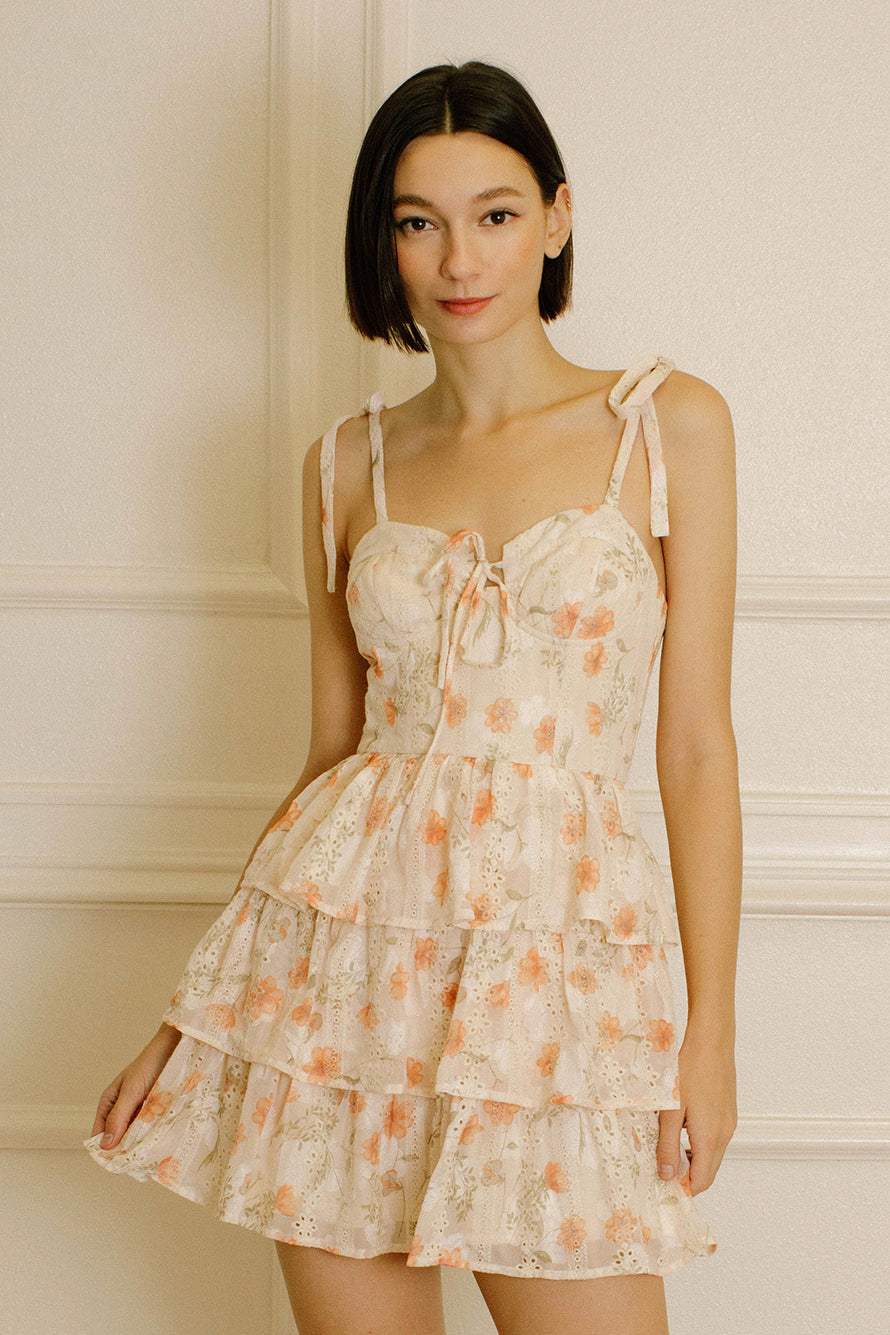 Woman wearing a peach dress with floral pattern, eyelet details and self-tie shoulder straps, against a neutral background