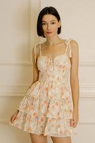 Woman wearing a peach dress with floral pattern, eyelet details and self-tie shoulder straps, against a neutral background