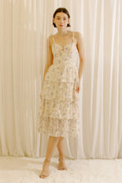 Woman wearing a cream floral midi dress with an eyelet design and a tiered skirt standing against a white curtain