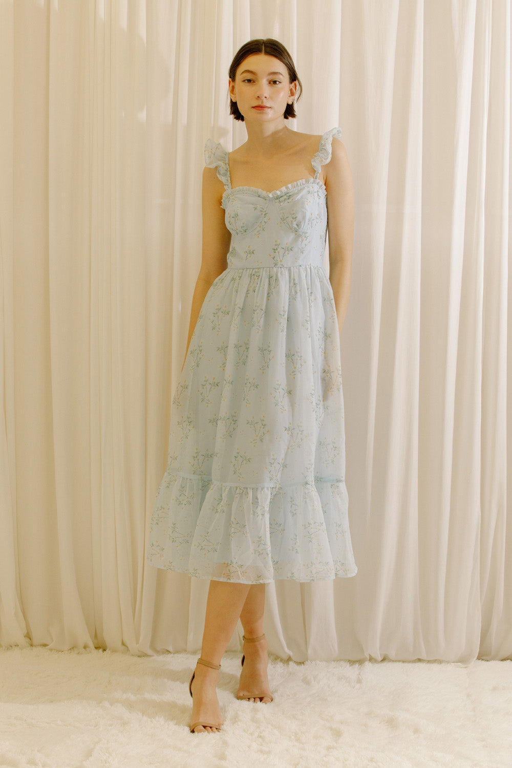 A woman wearing a light blue tiered midi dress with a floral print, standing in front of a cream-colored curtain backdrop.