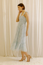 A woman wearing a light blue sleeveless midi dress with a delicate floral print, standing in front of a soft cream-colored curtain backdrop.