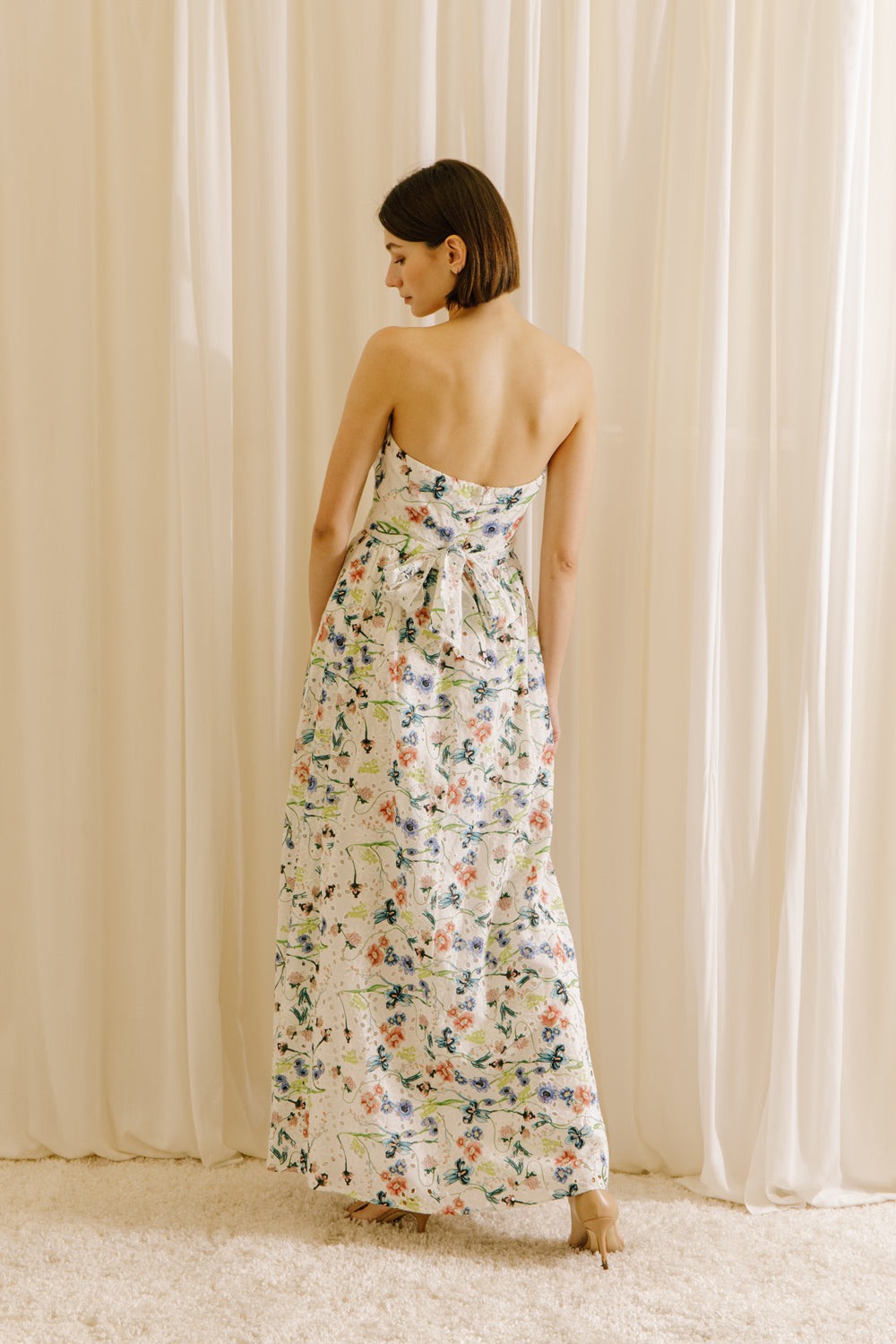 Back view of a woman wearing a blue floral strapless dress against a beige curtain background