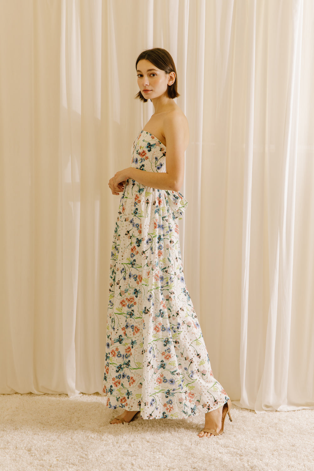 Side view of a woman wearing a blue floral strapless dress against a beige curtain background