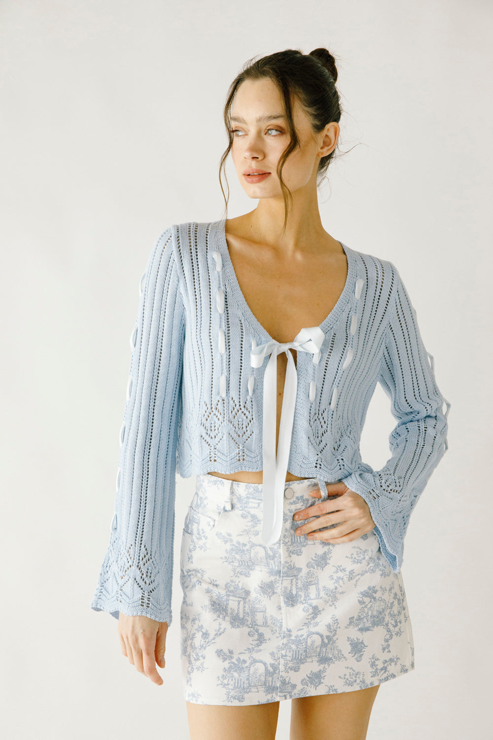 A woman wearing a blue knit cardigan with lace-up details and a toile print skirt on a white background
