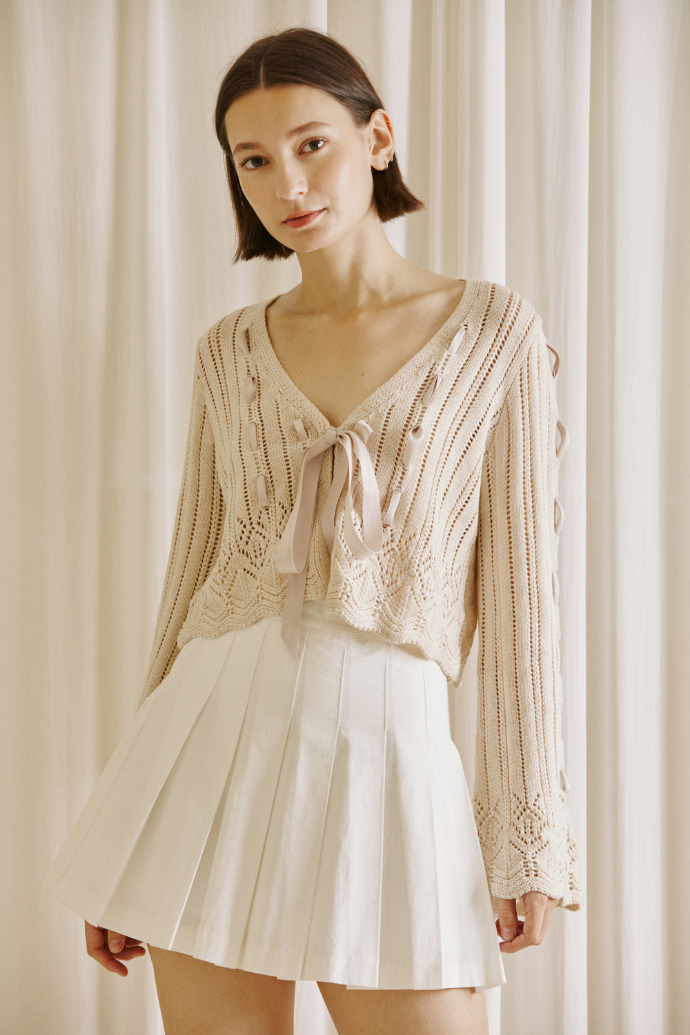 Woman wearing a beige knit cardigan with lace-up details and a white pleated skirt on a white background