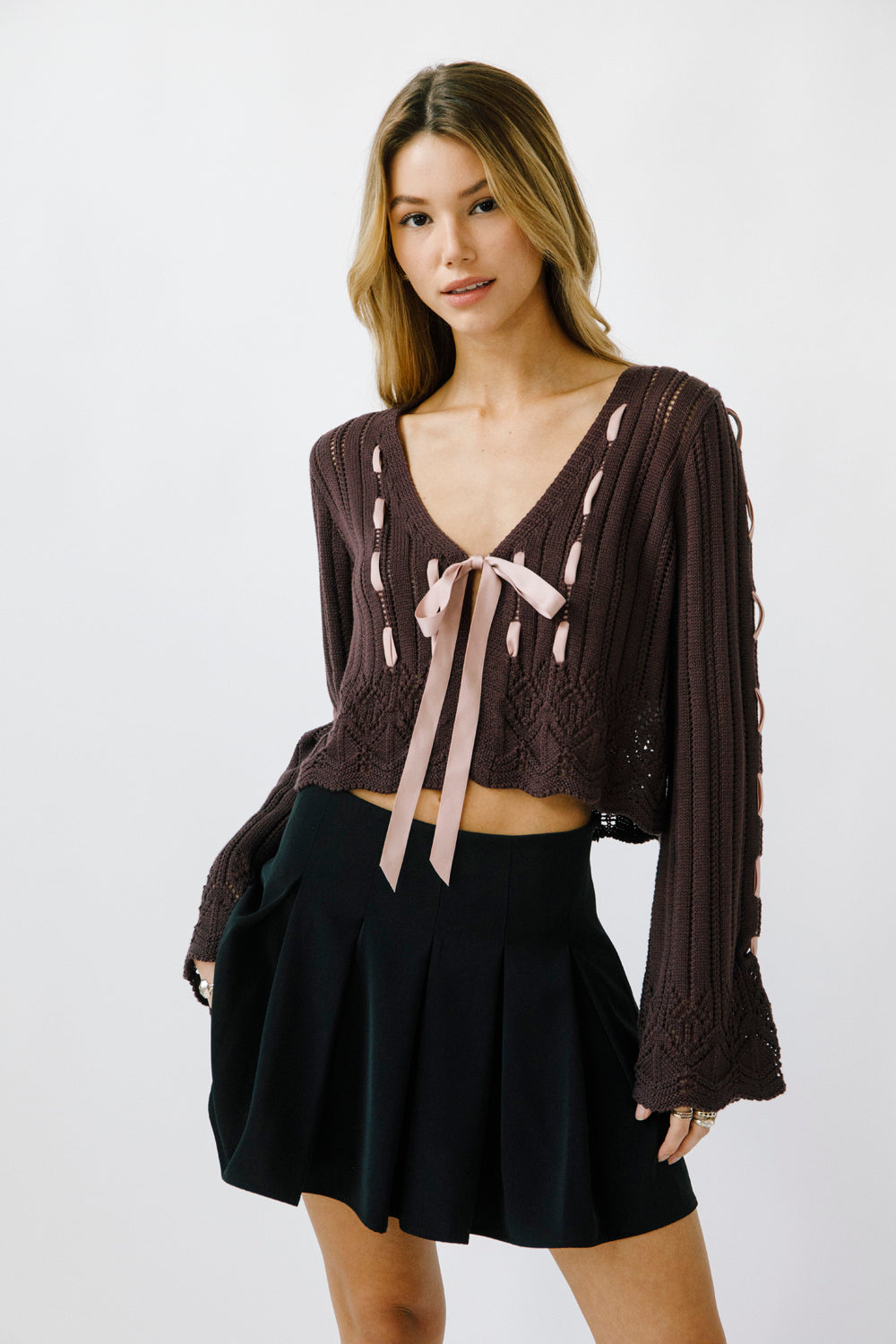 Woman wearing an eggplant knit cardigan with lace-up details and a black pleated skirt on a white background