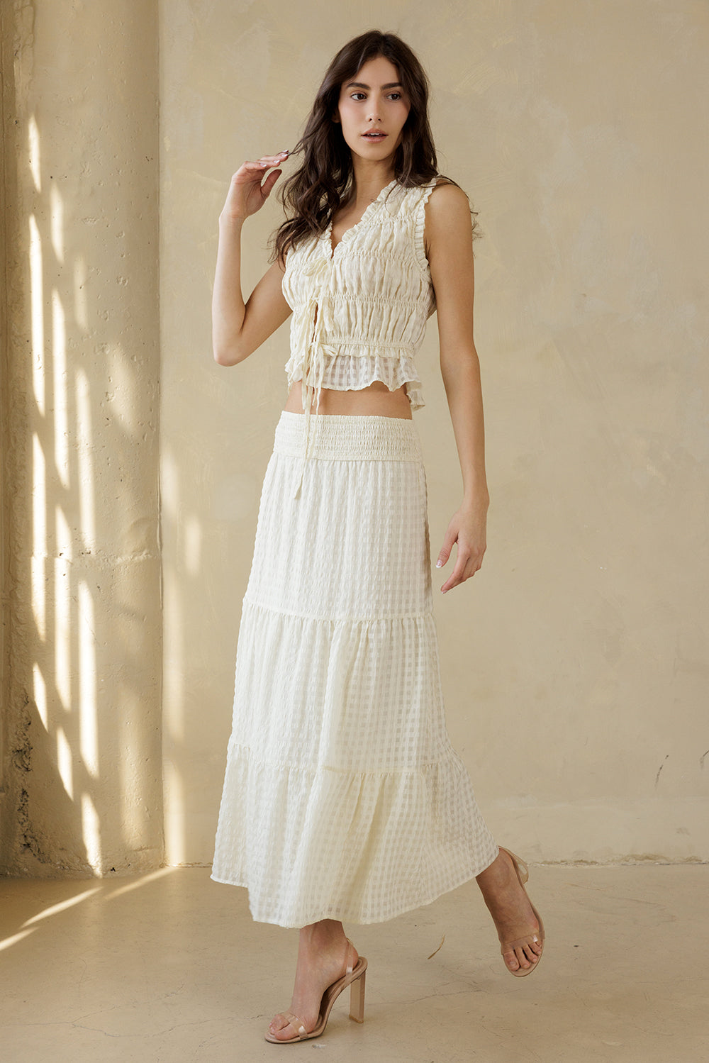 Woman wearing a cream textured maxi skirt with a subtle checkered pattern and a cream top standing against a beige wall with sunlight casting shadows.