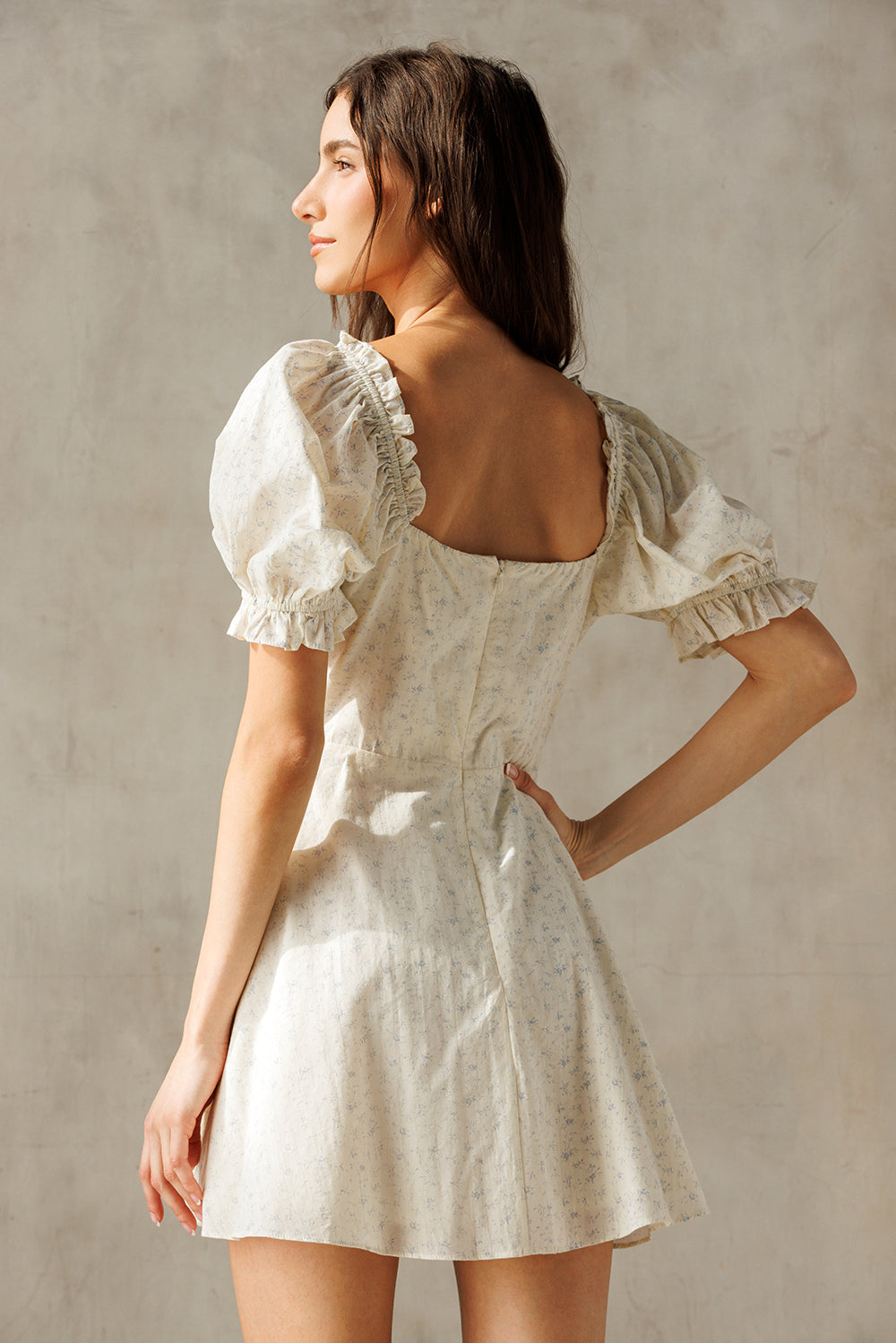 A woman viewed from behind wearing a short puff-sleeve dress with a square open back and subtle floral print, photographed against a textured neutral wall.