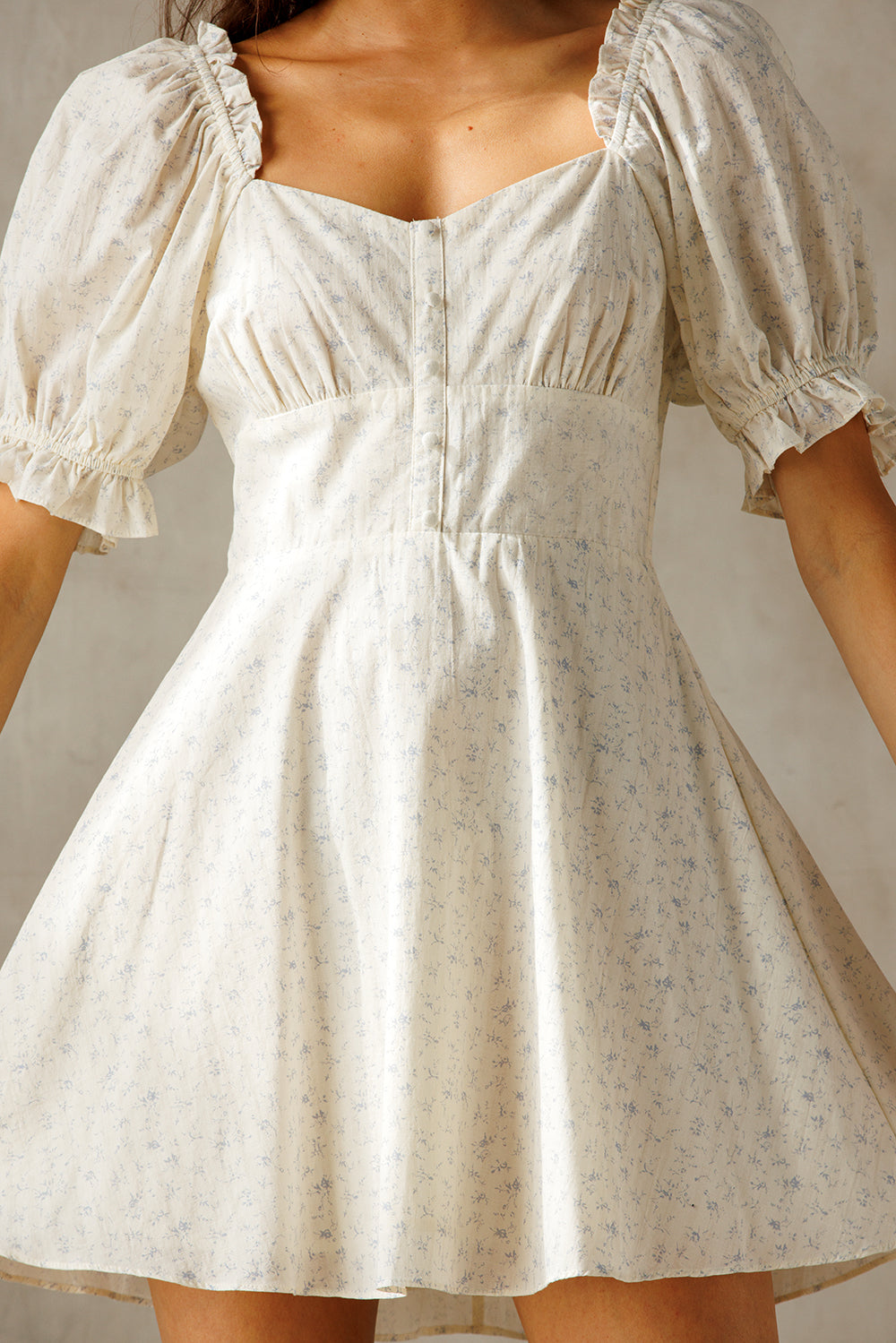 A woman wearing a short puff-sleeve dress with a square neckline and a flared skirt. The dress is cream with a subtle light blue floral print, photographed against a neutral light gray background.