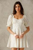A woman modeling a short puff-sleeve dress with a square neckline, gathered bodice, and flared skirt in a cream fabric with a subtle floral print, set against a textured neutral background.