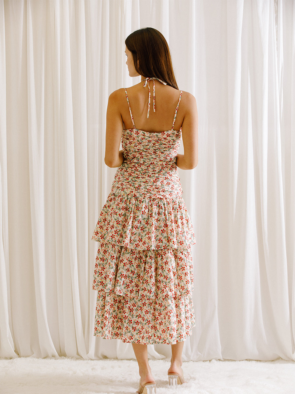 Back view of a woman wearing a floral dress with ruched upper body standing against a white curtain