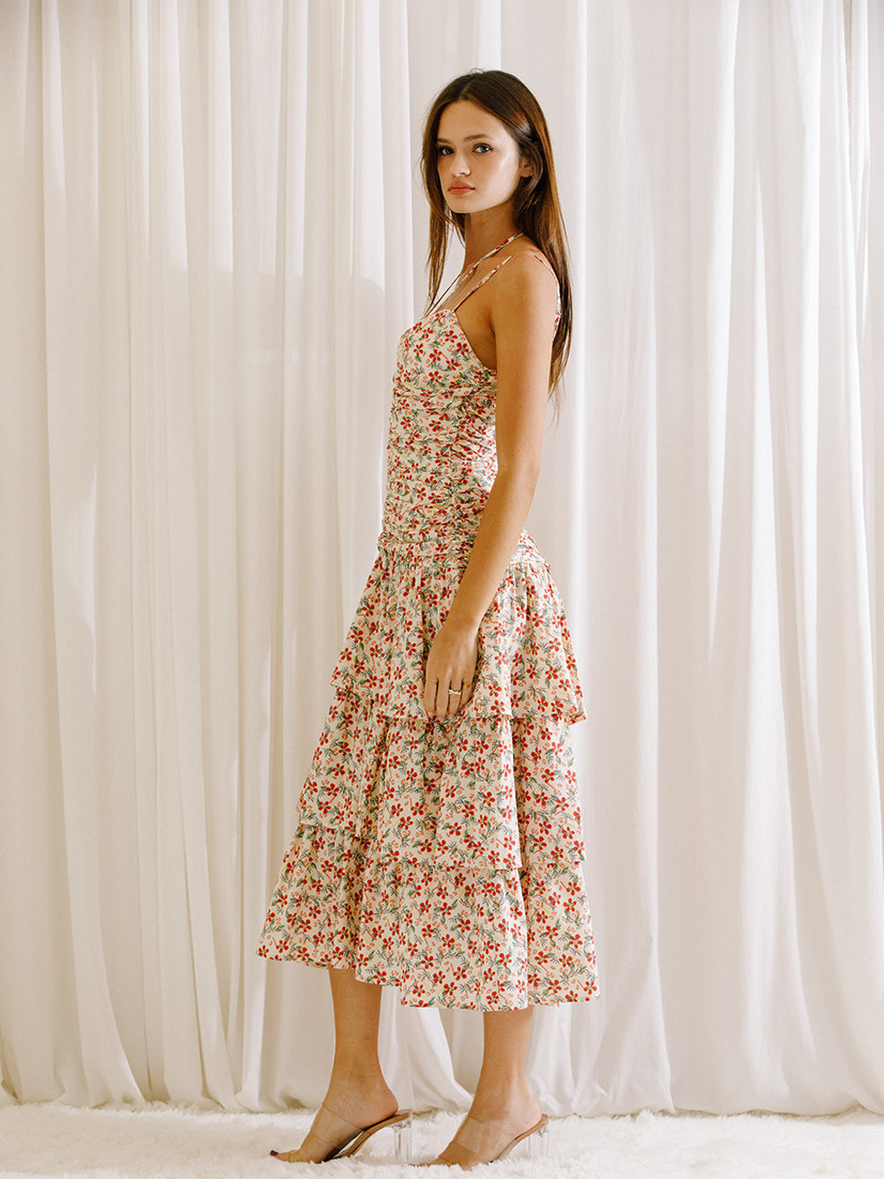 Side view of a woman wearing a floral midi dress with ruched upper body standing against a white curtain