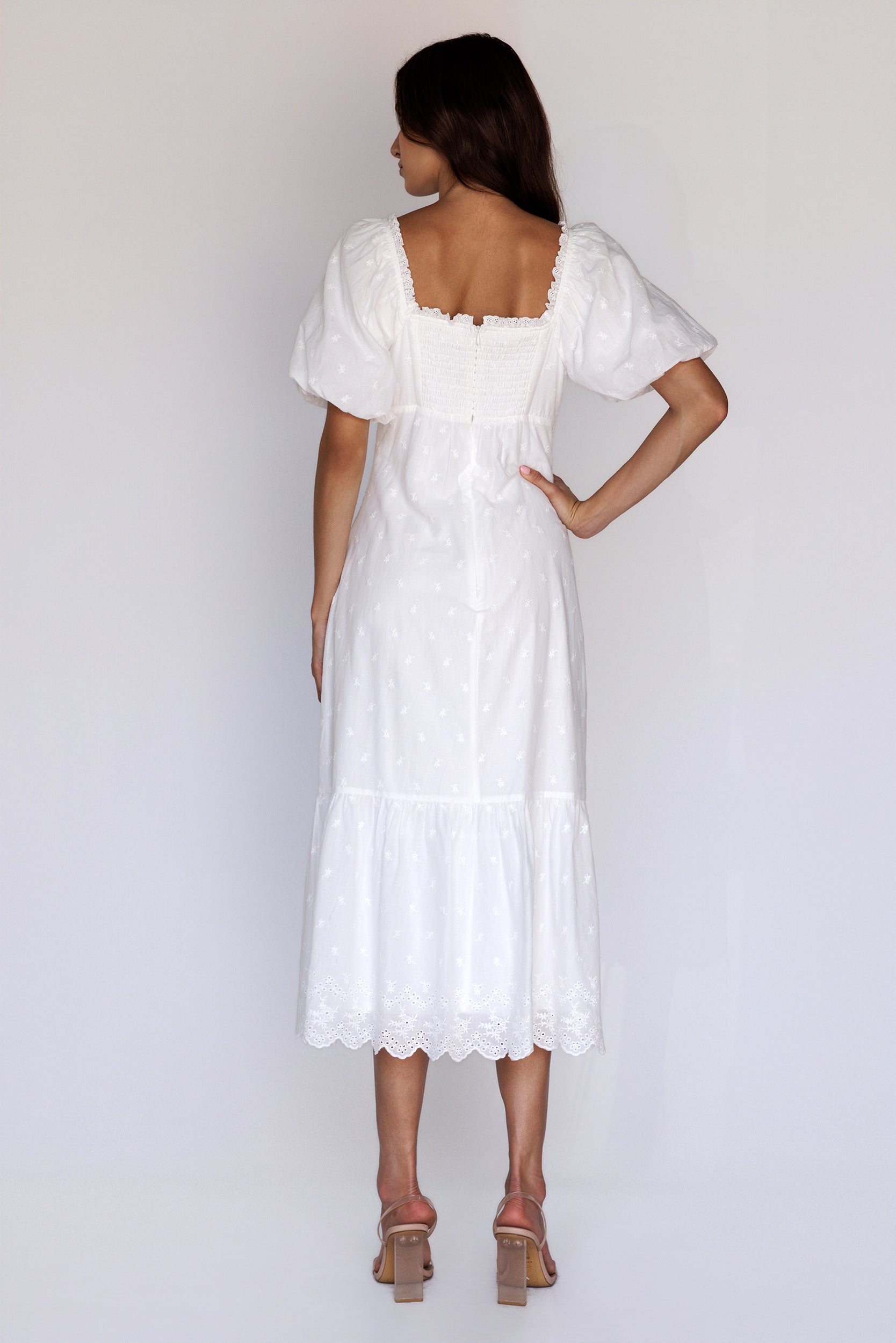 A woman viewed from behind wearing a timeless white embroidered midi dress with puff sleeves, a smocked back, a back invisible zipper and scalloped eyelet hem.