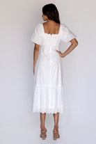 A woman viewed from behind wearing a timeless white embroidered midi dress with puff sleeves, a smocked back, a back invisible zipper and scalloped eyelet hem.