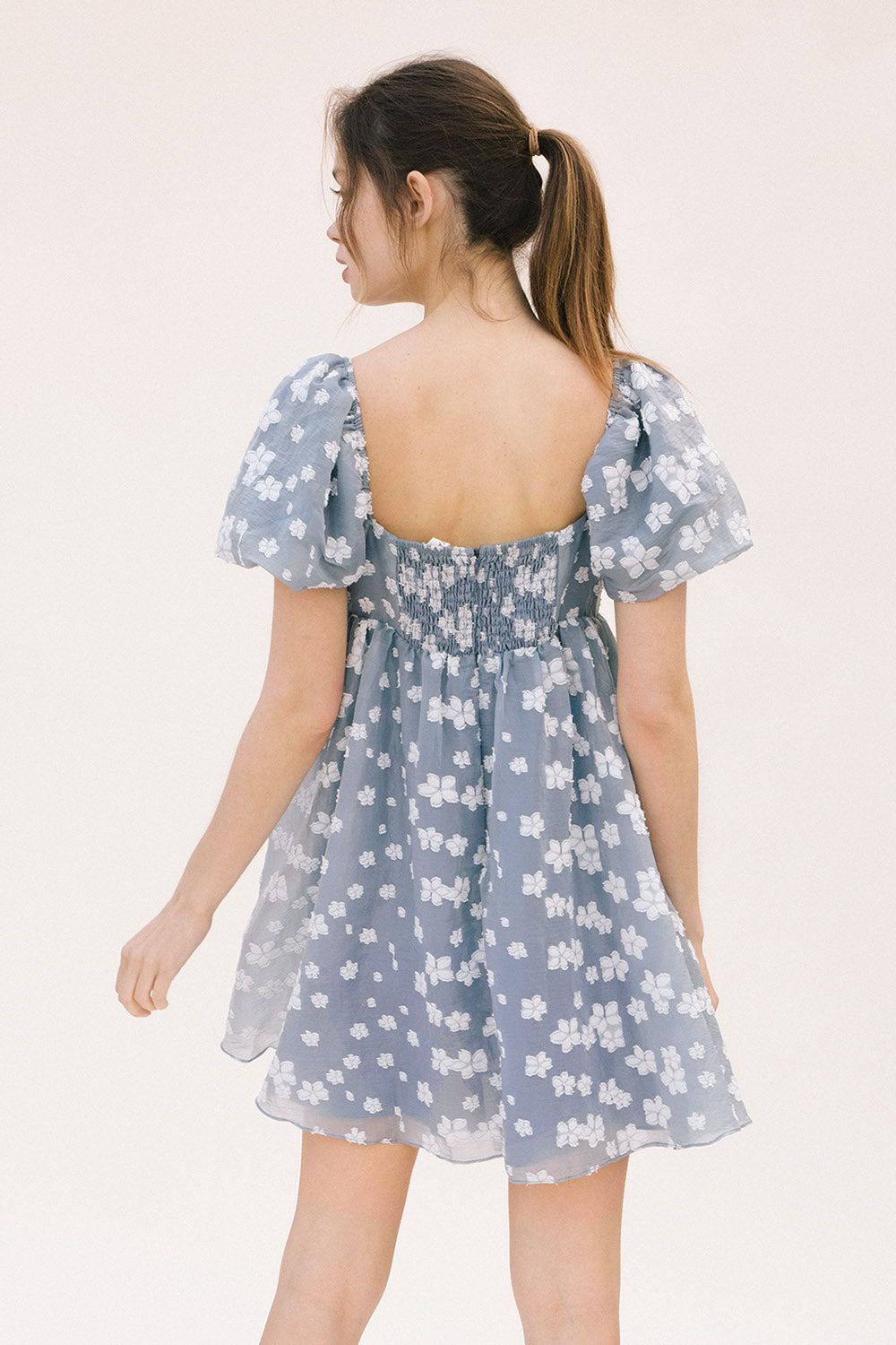 Back view of a woman wearing a blue dress with white floral embroidery, puff sleeves, back invisible zipper and smocked upper back on a white background