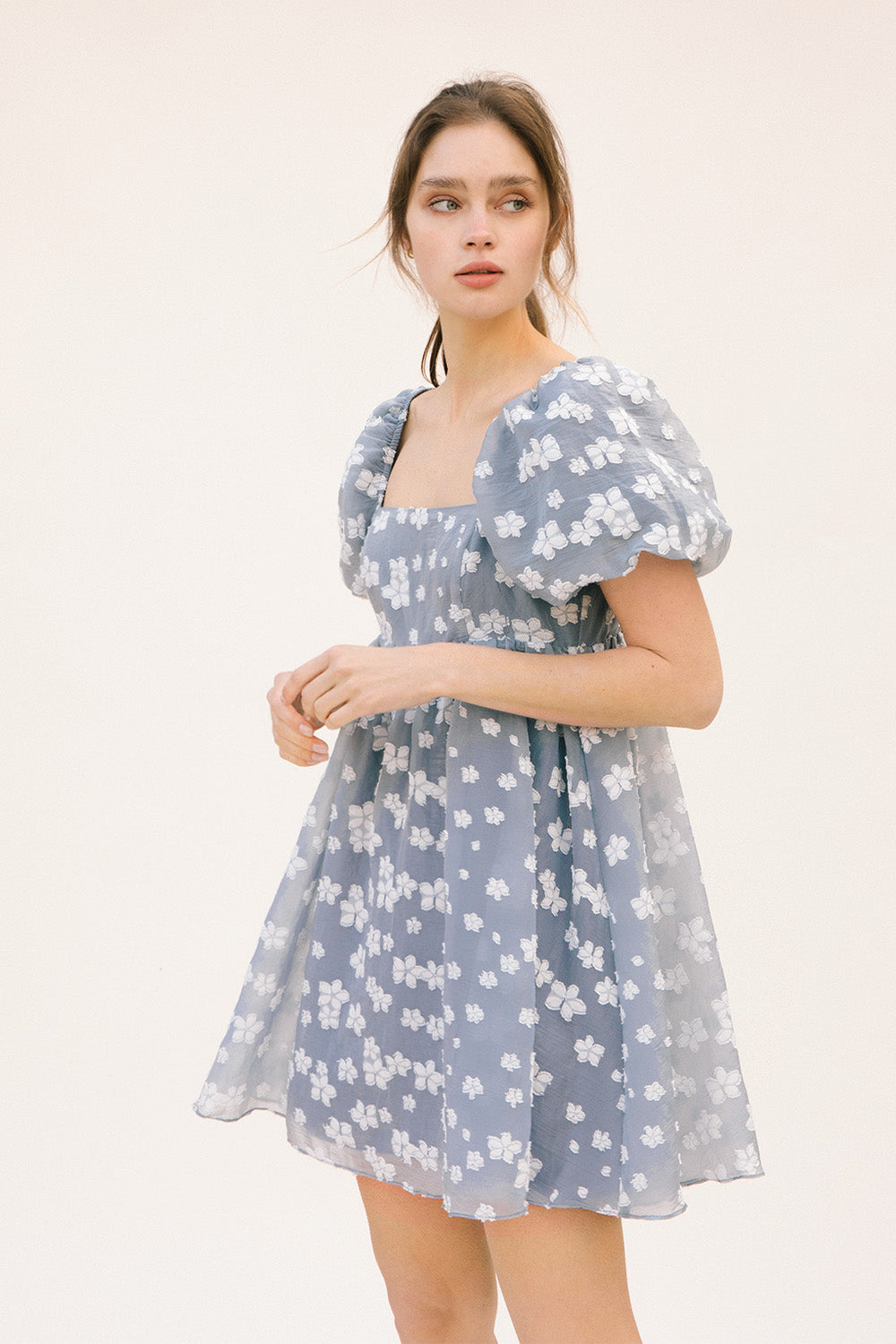 Side view of a woman wearing a blue dress with white floral embroidery and puff sleeves on a white background