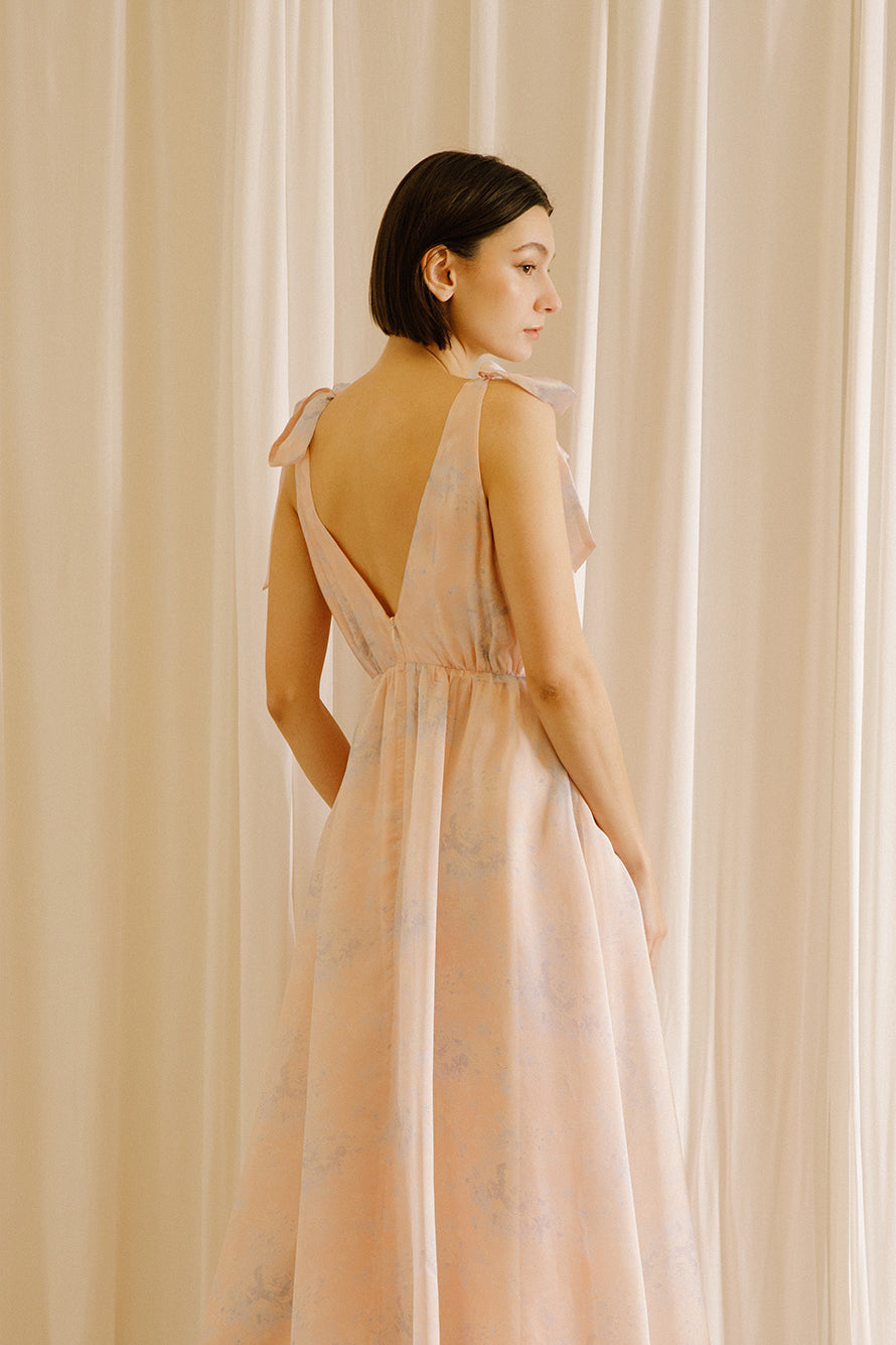 Back view of a woman wearing a light pink dress with thin straps against a beige curtain background