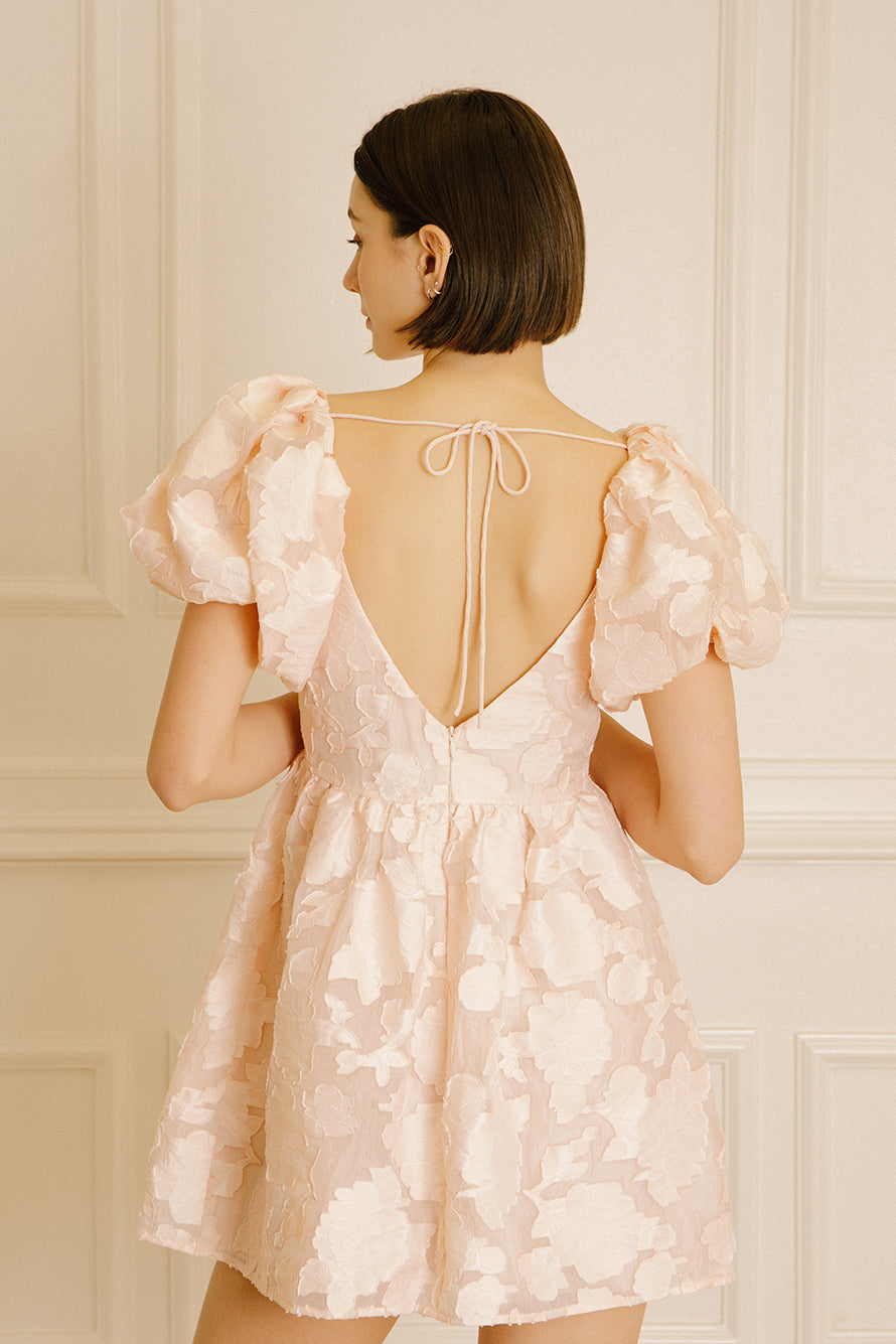 Back view of a woman wearing a peach dress with an invisible back zipper and a thin self-tie strap against a neutral wall.
