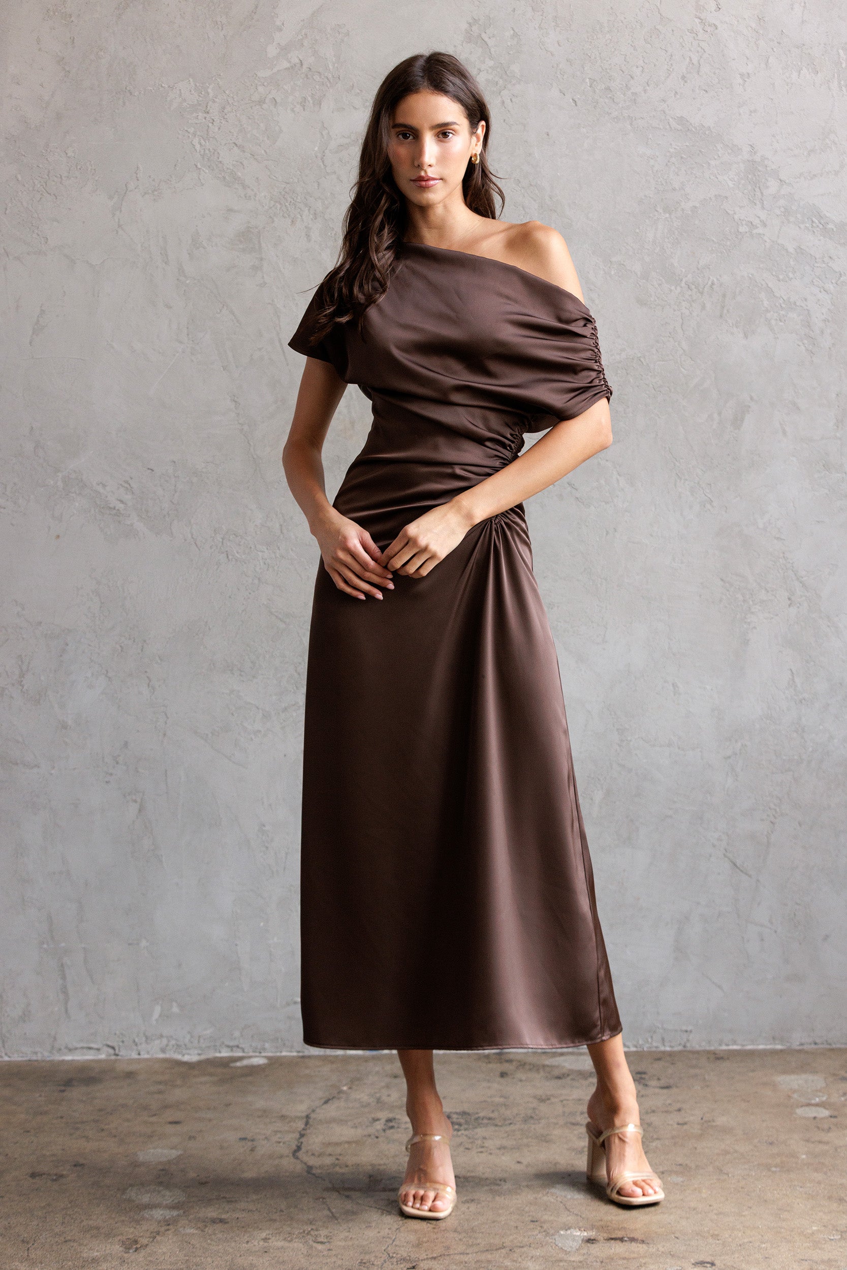 Woman wearing a brown off-shoulder dress against a gray concrete wall.