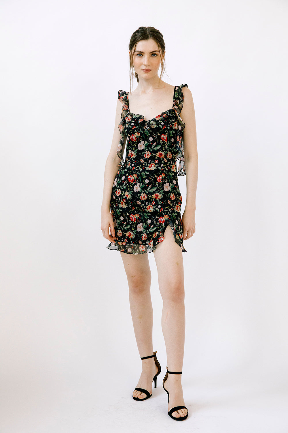 Woman wearing a black flowy mini dress with a floral pattern and black high-heels on a white background