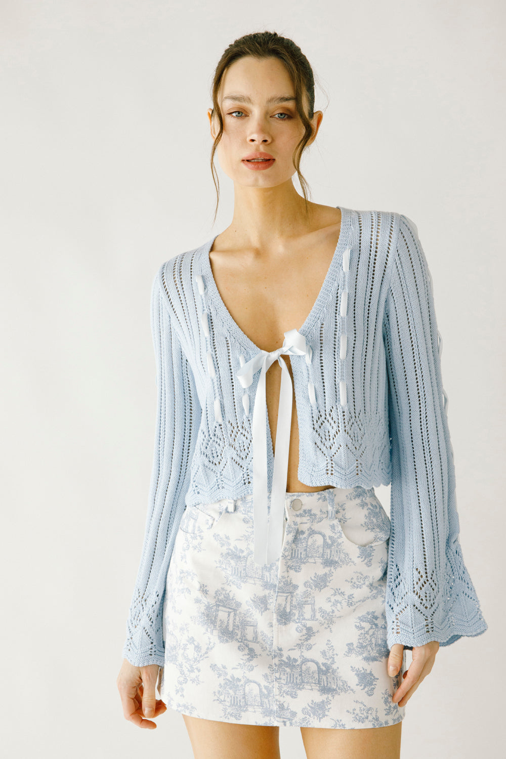 Woman wearing a light blue crochet cardigan and a blue and white skirt on a plain background